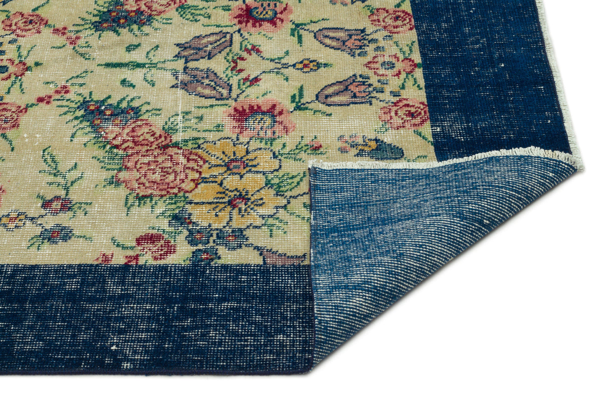 Beige rug with a floral pattern in red, yellow, and green, featuring a wide navy blue border and a folded corner.