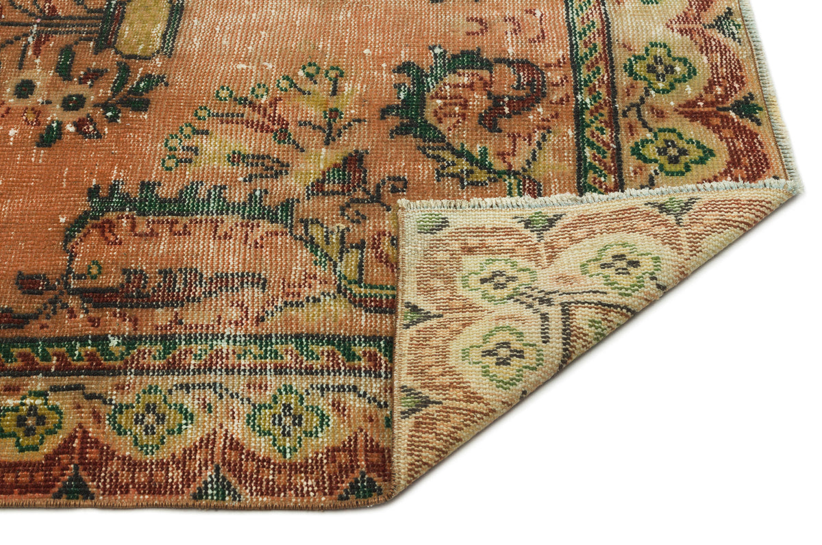 Distressed terracotta rug with green and cream floral motifs, showing a folded corner on a white background.