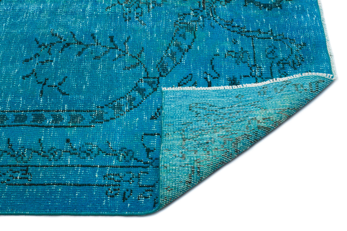 Bright turquoise rug with a distressed black floral medallion pattern, shown with a corner folded over on white.