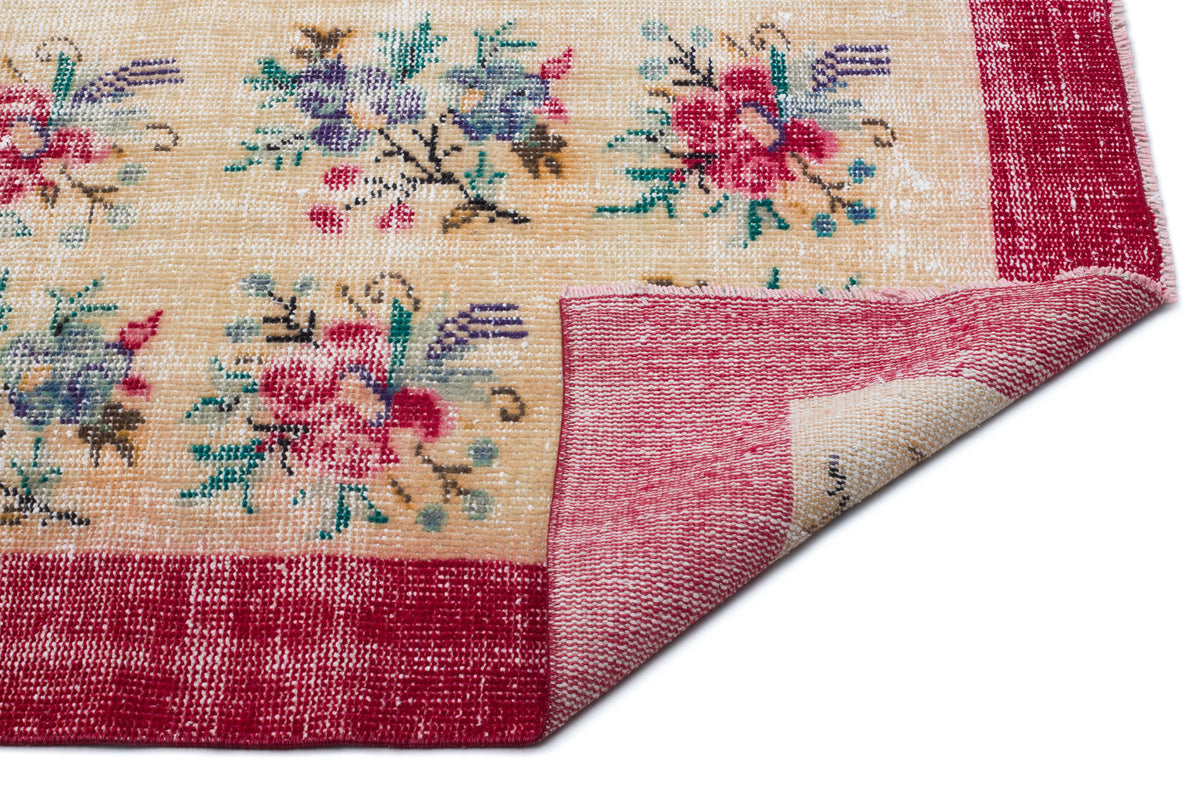 Beige rug with a distressed floral pattern in red, blue, and green, featuring a wide red border and a folded corner.