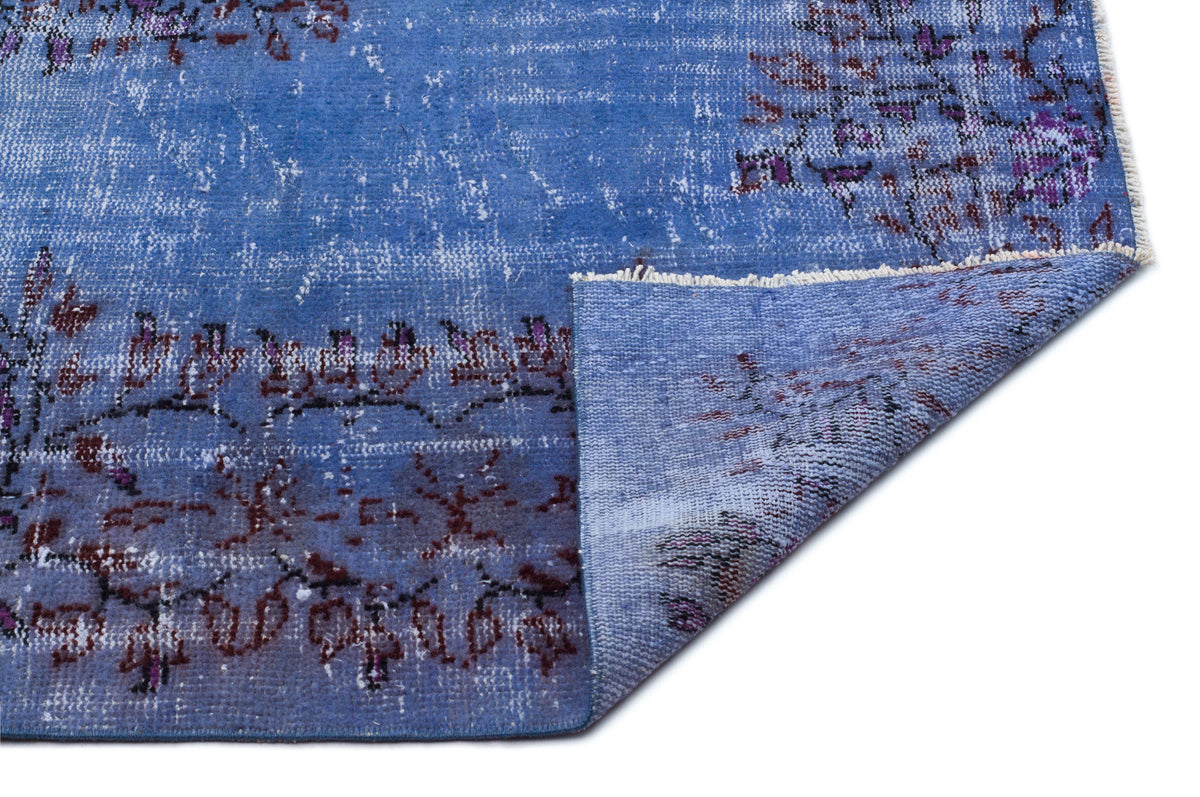 Distressed blue rug with purple and black floral motifs, featuring a folded corner revealing the woven backing.