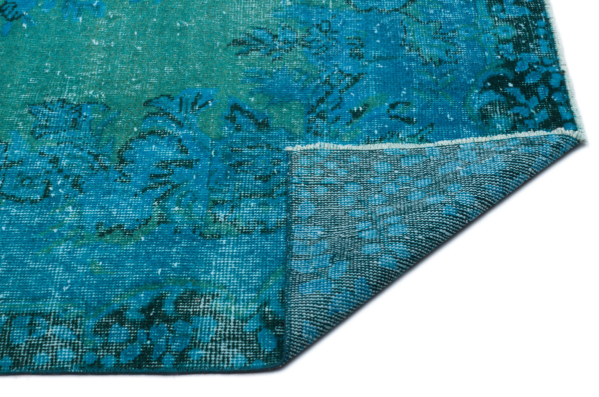 Teal and blue distressed rug with a faded floral medallion pattern, shown with a corner folded over on a white background.