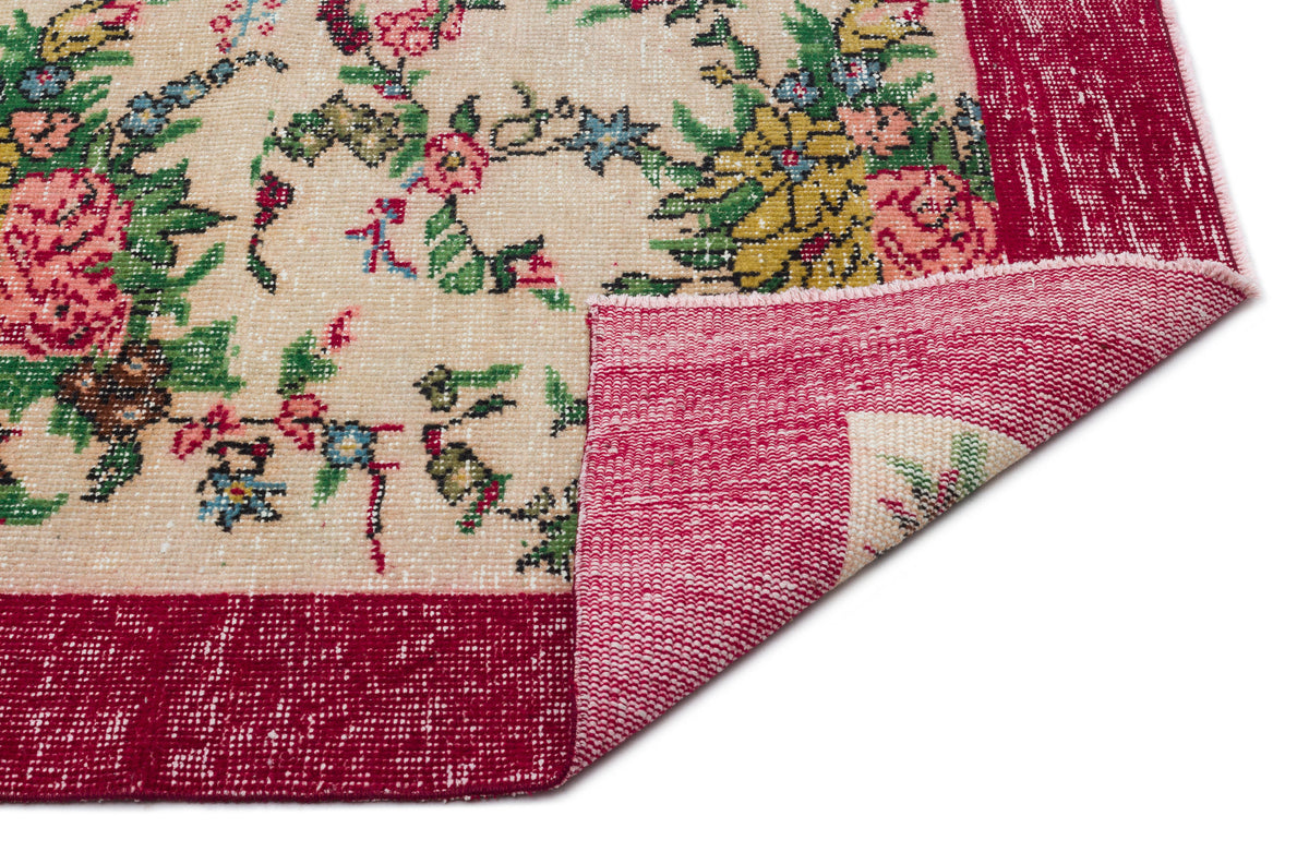 Beige rug with a floral pattern in pink, green, and blue, featuring a wide red border and a folded corner.