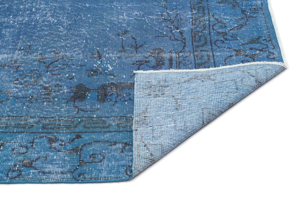 Blue distressed rug with a dark grey geometric border and floral motifs, shown with a corner folded over on white.