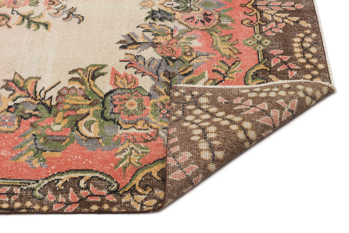 Beige rug with a floral medallion pattern in coral, green, and brown, featuring a folded corner showing the woven backing.