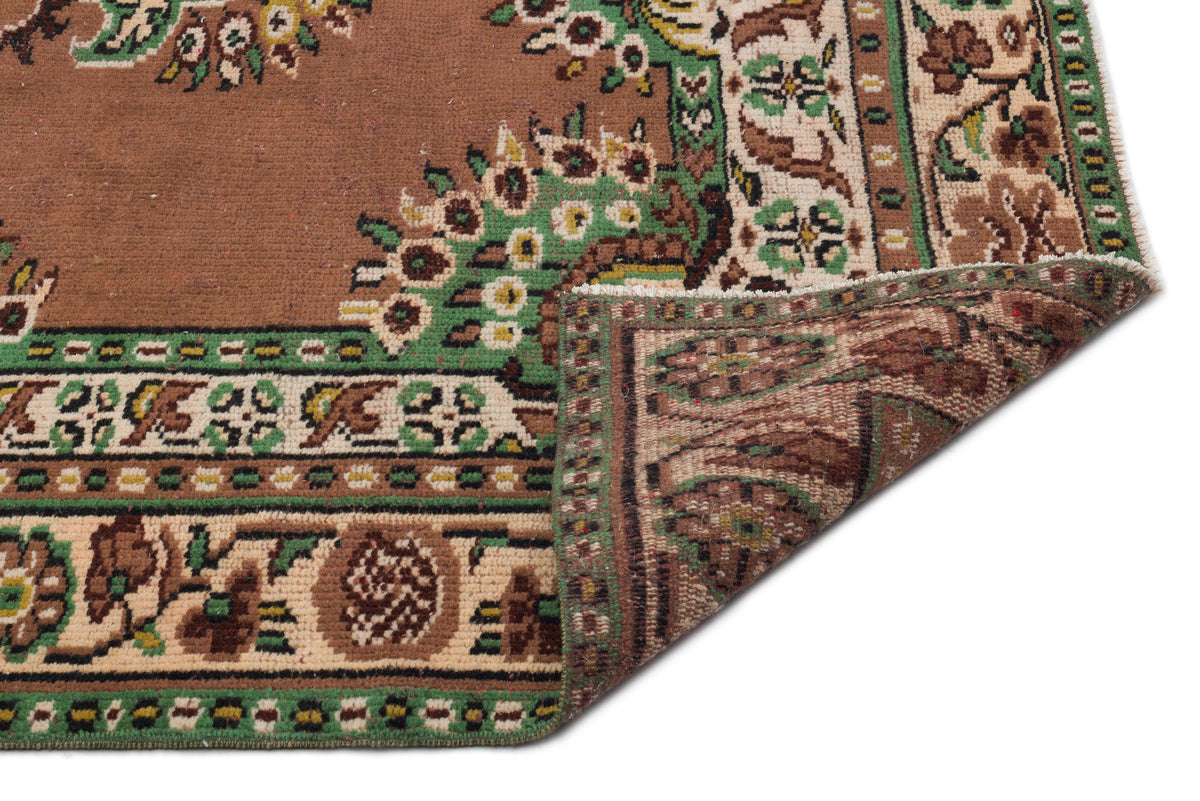 Brown traditional rug with green and cream floral border patterns, shown with a folded corner on a white background.