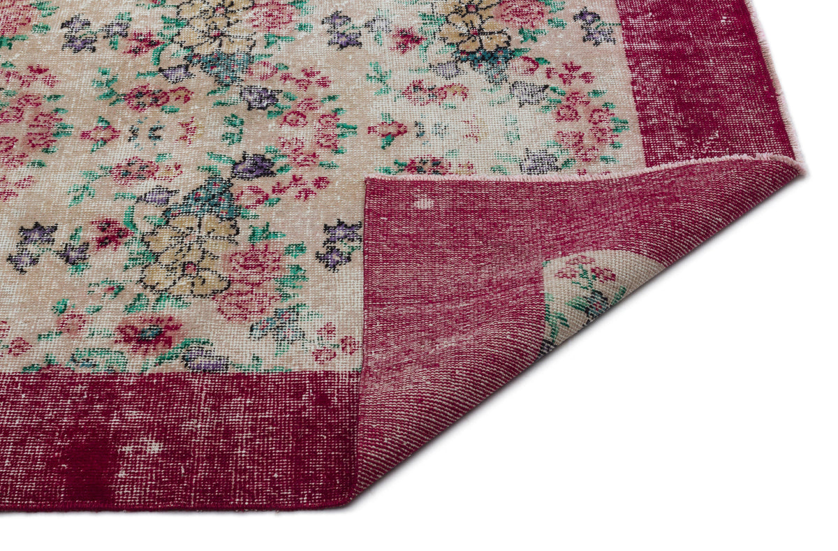 Beige rug with a distressed floral pattern in pink, green, and purple, featuring a solid magenta border and folded corner.