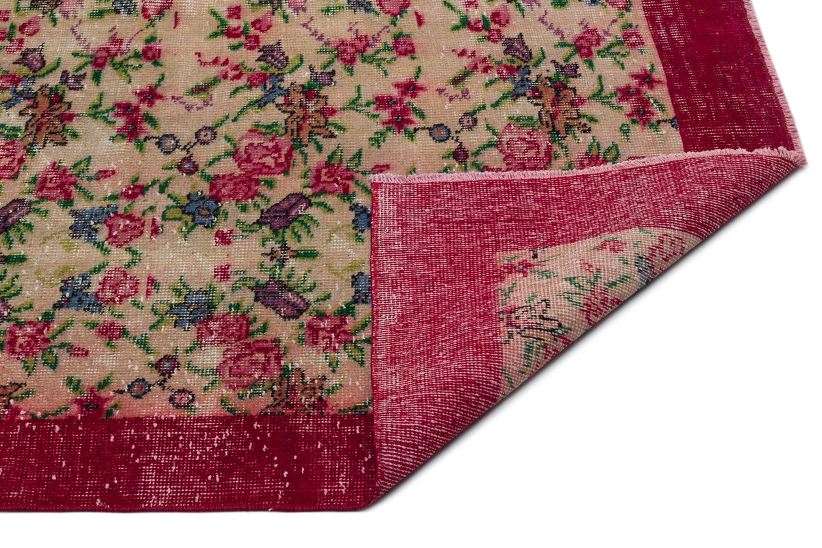 Beige rug with a distressed floral pattern in pink, green, and blue, featuring a wide red border and a folded corner.