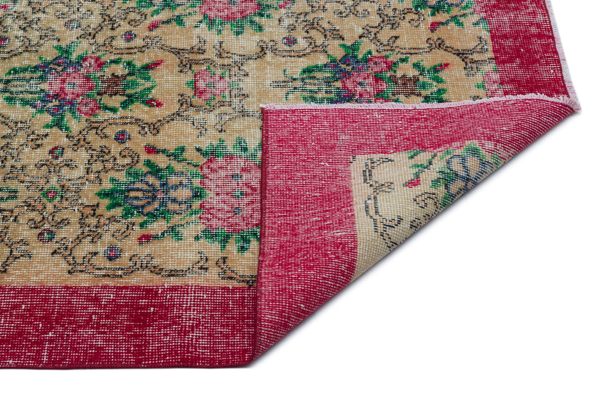 Beige rug with a floral pattern in pink, green, and blue, featuring a wide red border and a folded corner.