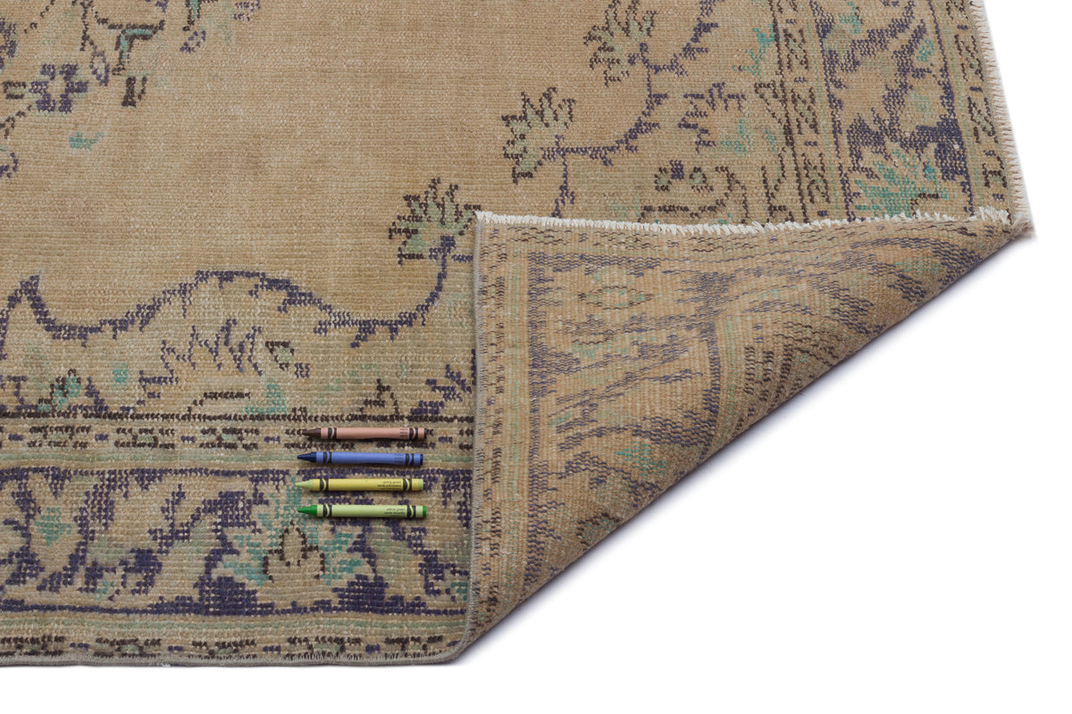 Tan vintage-style rug with a distressed floral pattern in blue and green, shown with a folded corner and four crayons.