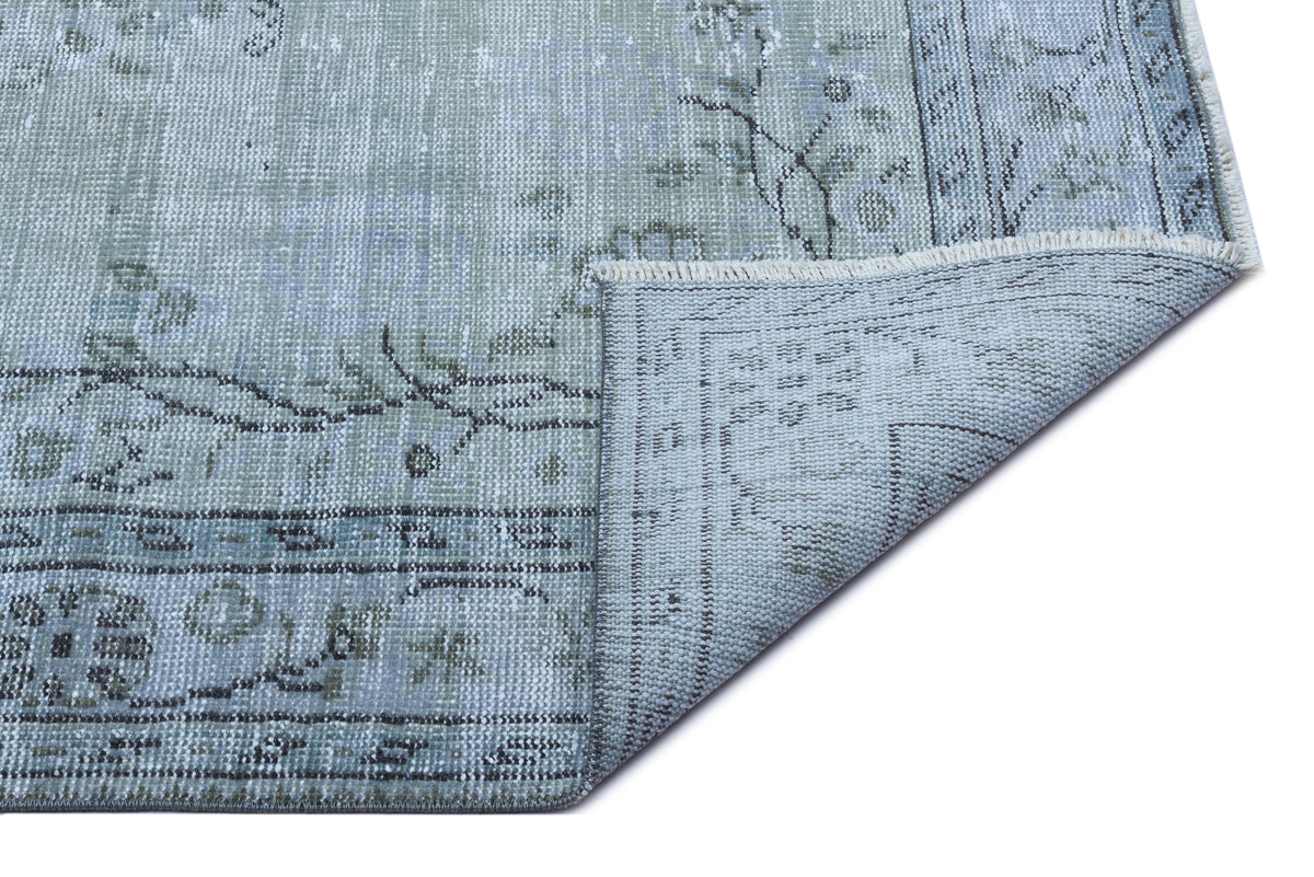 Distressed sage green and blue traditional rug with a floral medallion pattern and a folded corner showing the backing.