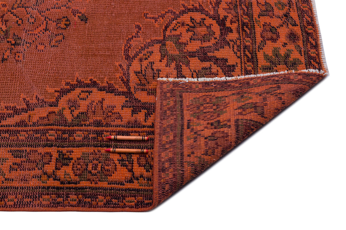 Rust-colored vintage-style rug with a dark floral border and a corner folded over, isolated on a white background.