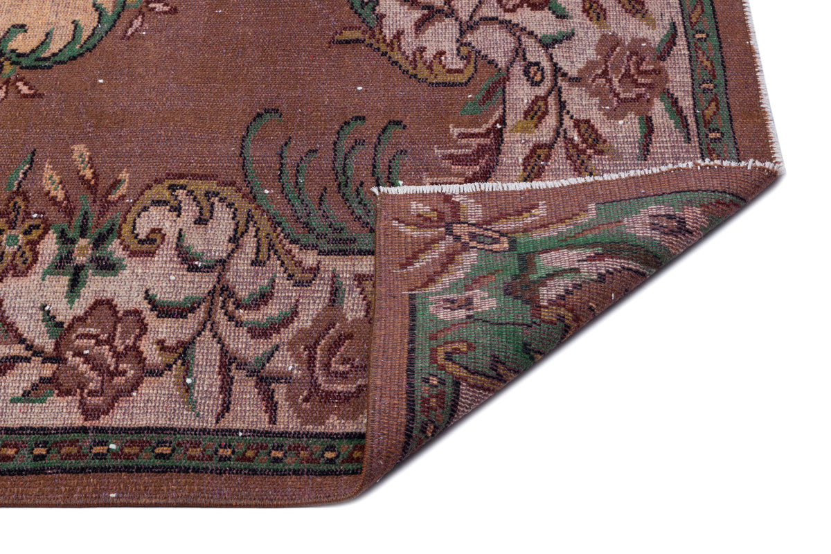 Brown traditional rug with a floral pattern in green and beige, shown with a corner folded over on a white background.