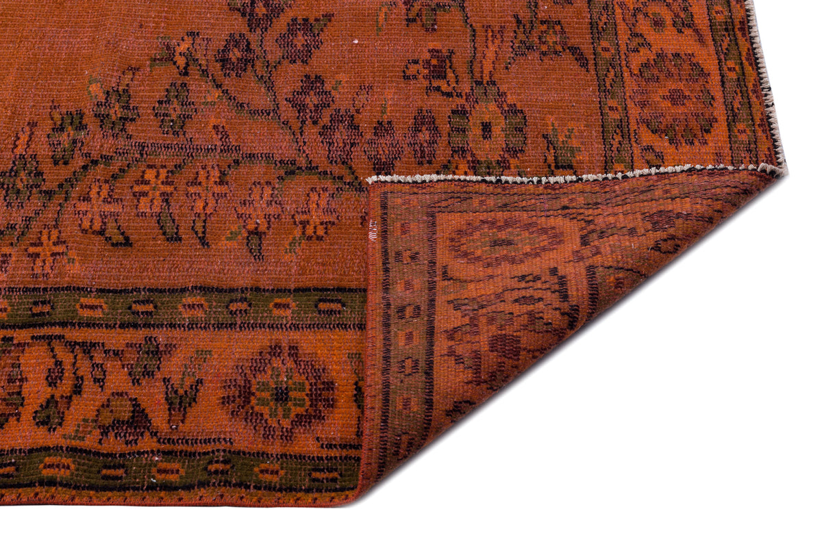 Rust-colored rug with a distressed floral medallion pattern and dark green border, shown with a folded corner.
