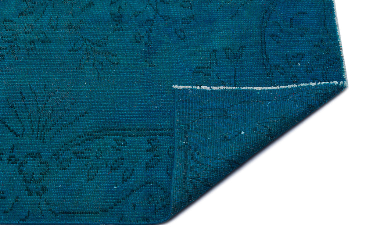 Teal rug with a subtle, dark floral pattern and a folded corner revealing the woven texture on a white background.