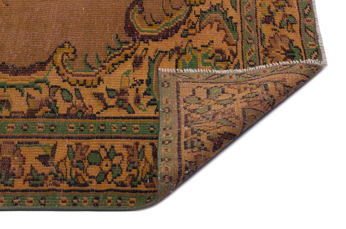 Brown and green vintage-style rug with a floral medallion border, shown with a corner folded over on a white background.