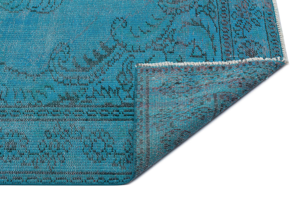Bright turquoise rug with a distressed black floral medallion pattern and a folded corner showing the underside.