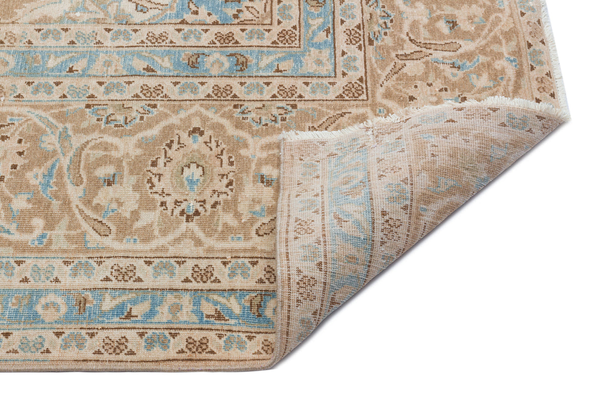Tan and light blue traditional rug with a floral medallion pattern and a folded corner showing the underside.
