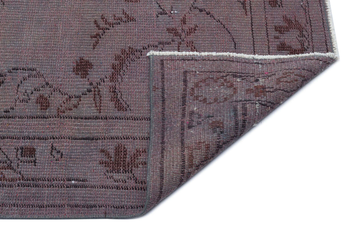 Muted purple rug with a dark brown floral pattern and a folded corner revealing the underside on a white background.