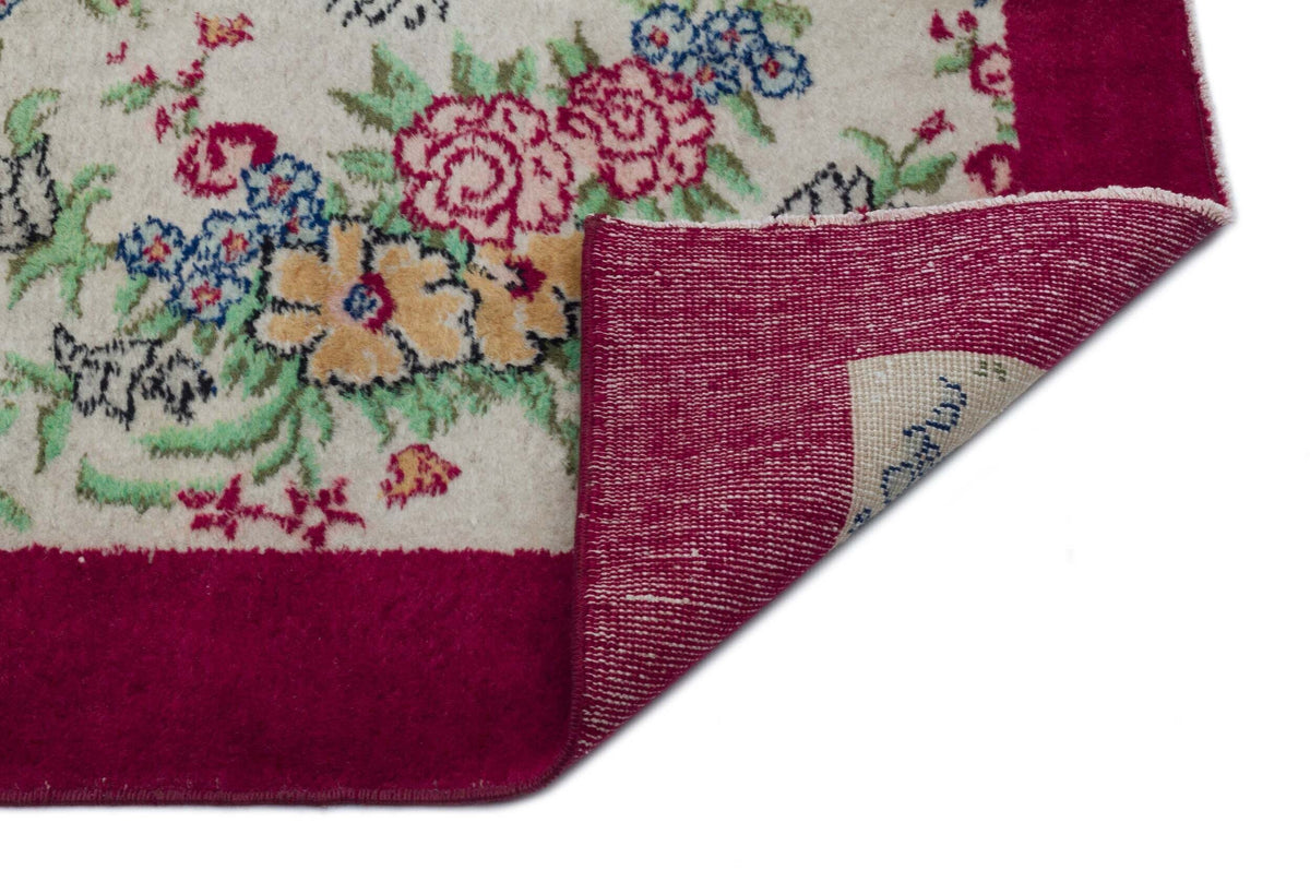 Cream rug with a floral pattern in pink, blue, and green, featuring a wide magenta border and a folded corner.