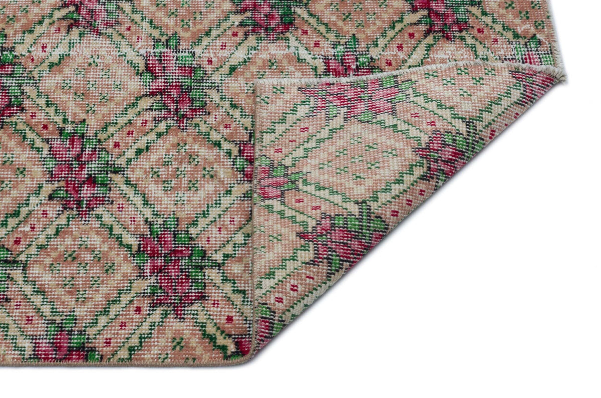 Beige, pink, and green vintage-style rug with a geometric floral pattern, shown with a folded corner on a white background.