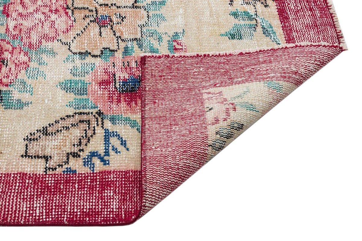 Beige rug with a floral pattern in pink and teal, featuring a folded corner revealing the red woven backing.