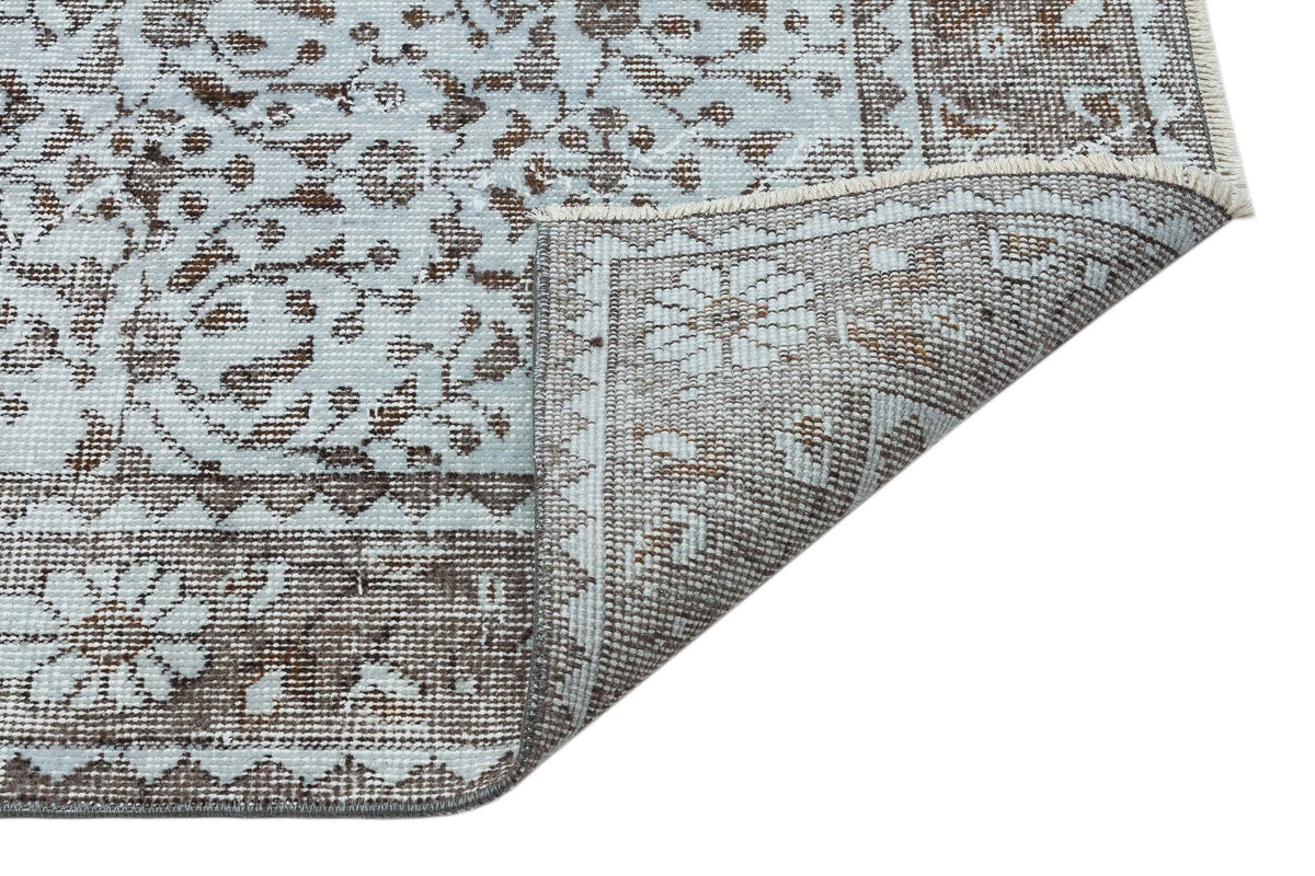 Light blue and brown rug with a traditional floral medallion pattern, shown with a corner folded over on white.