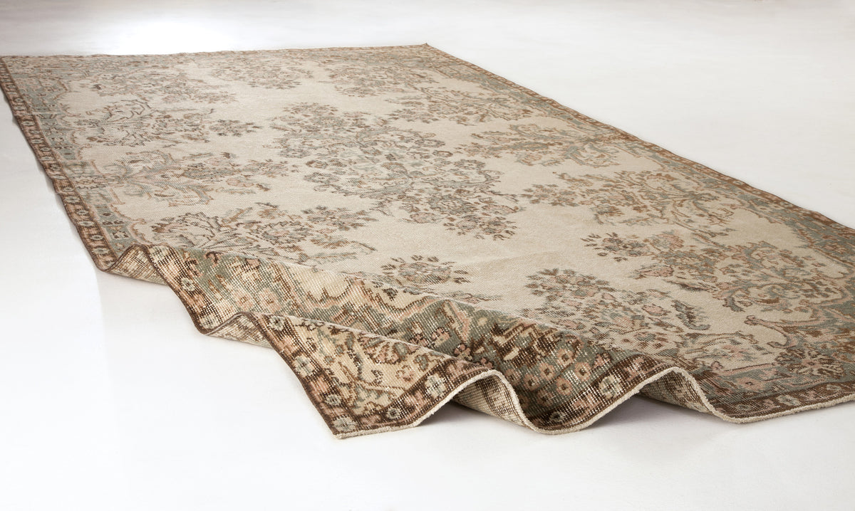 Beige and sage green vintage-style rug with a faded floral medallion pattern and a folded corner on a white background.