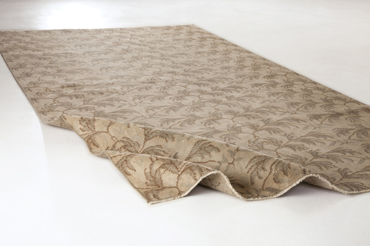 Beige and taupe rug with a repeating botanical leaf pattern, shown with one corner folded on a white background.