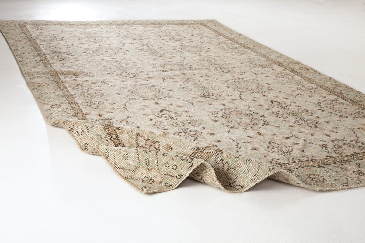 Beige and cream vintage-style rug with a faded floral medallion pattern and a folded corner on a white background.