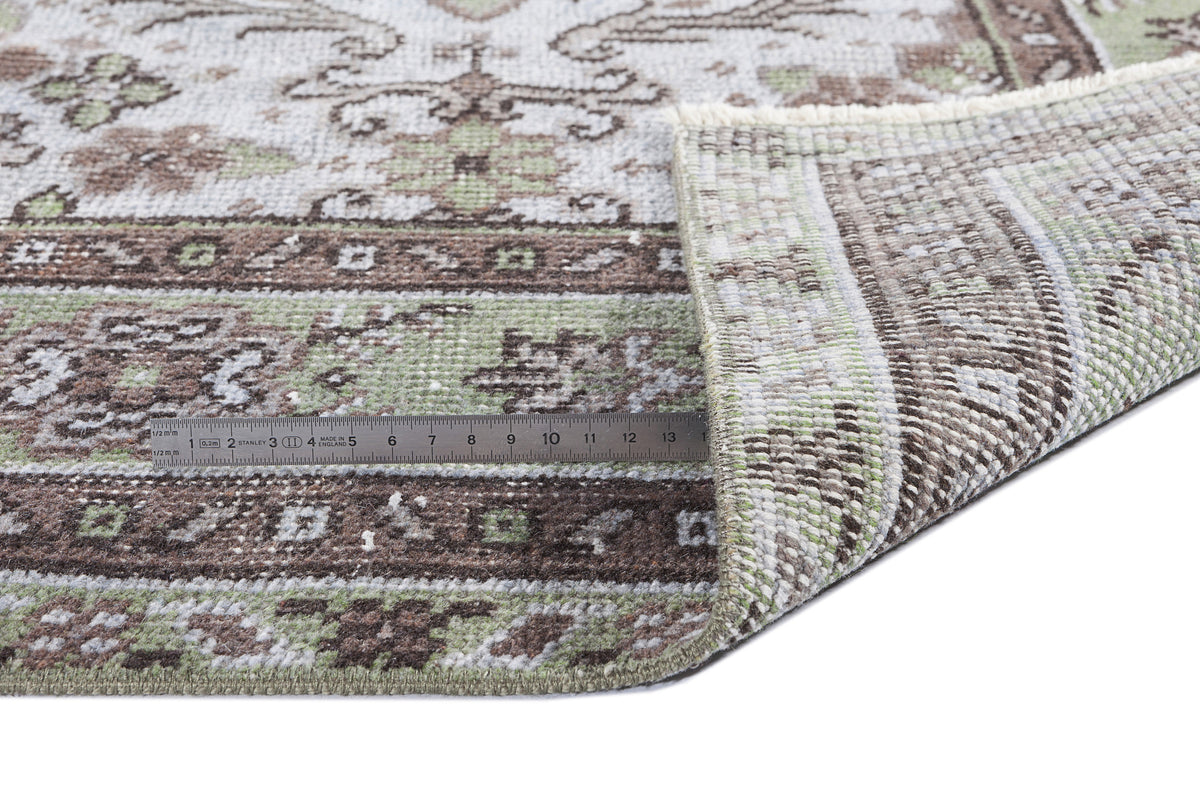 Distressed sage green and brown traditional rug with a folded corner and a metal ruler for scale on a white background.