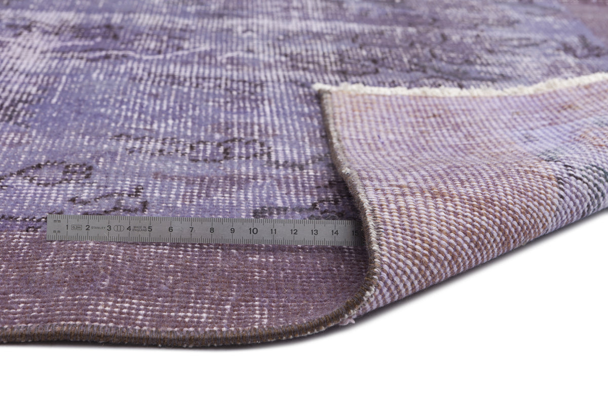 Distressed purple and grey vintage-style rug with a folded corner and a metal ruler for scale on a white background.