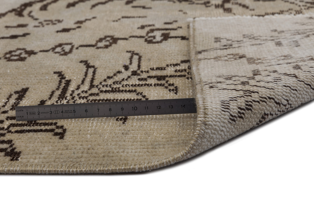 Beige rug with a dark brown abstract pattern, shown folded to reveal the backing with a metal ruler for scale.