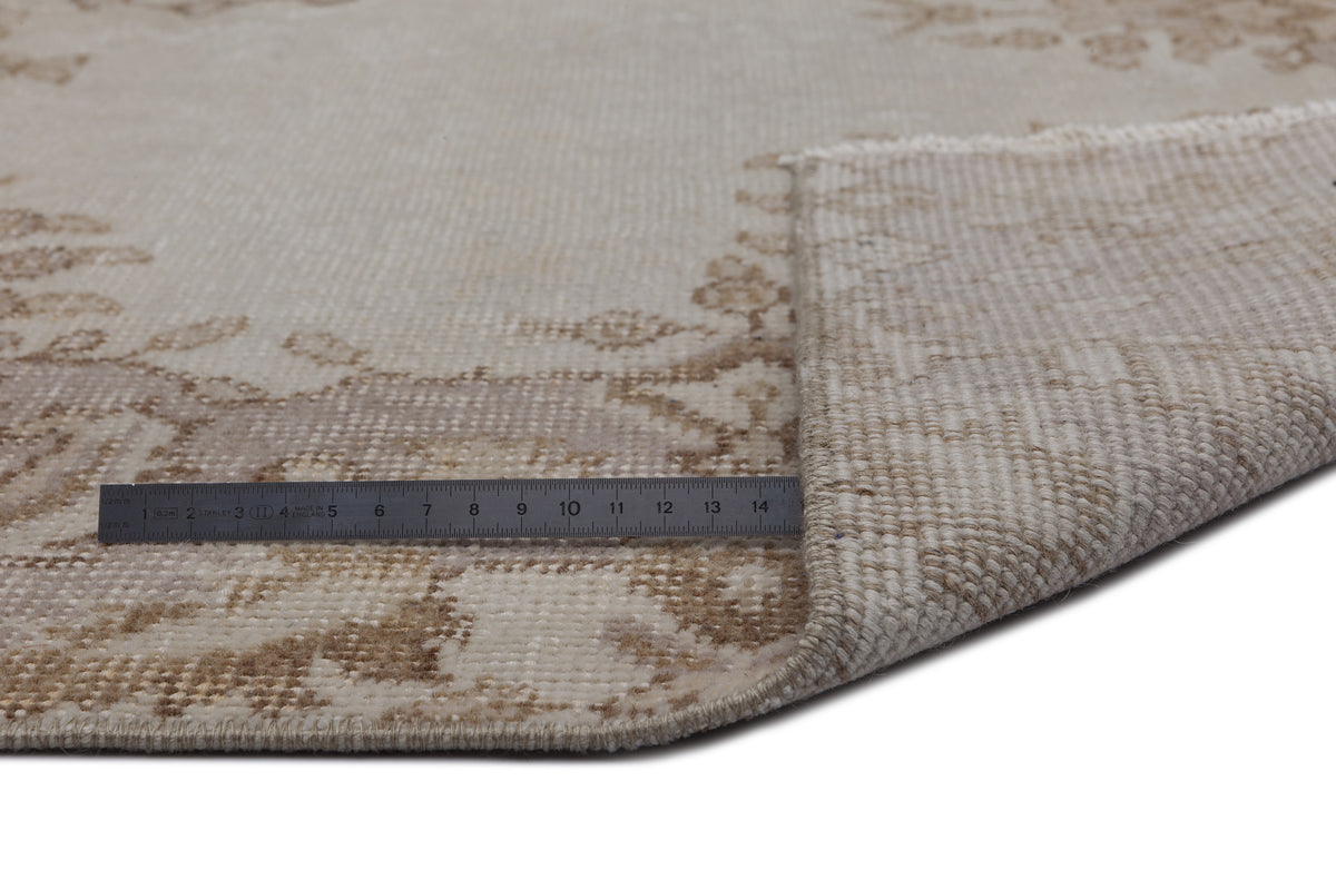Beige and cream vintage-style rug with a faded floral medallion pattern, shown with a corner folded and a metal ruler.