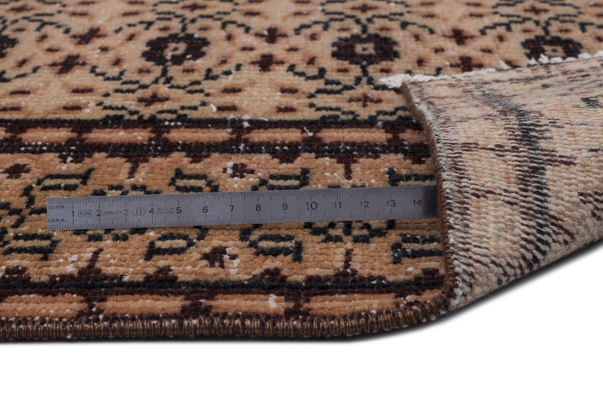 Tan and dark brown traditional patterned rug with a folded corner and a metal ruler showing pile thickness.