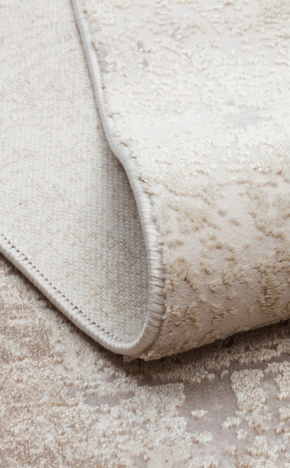 Beige and cream textured rug with a subtle distressed pattern, showing the woven backing and serged edge.