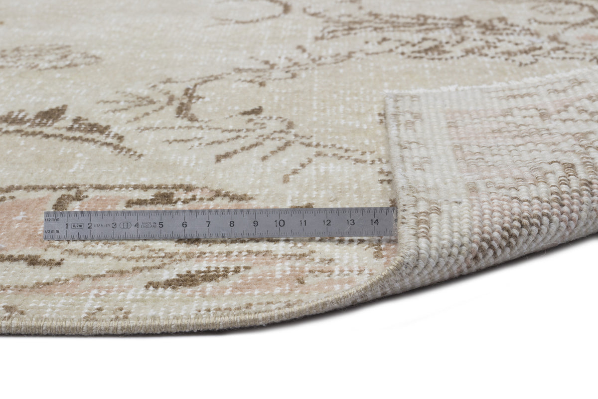 Beige and cream round rug with a distressed floral pattern, shown with a metal ruler for scale on a white background.