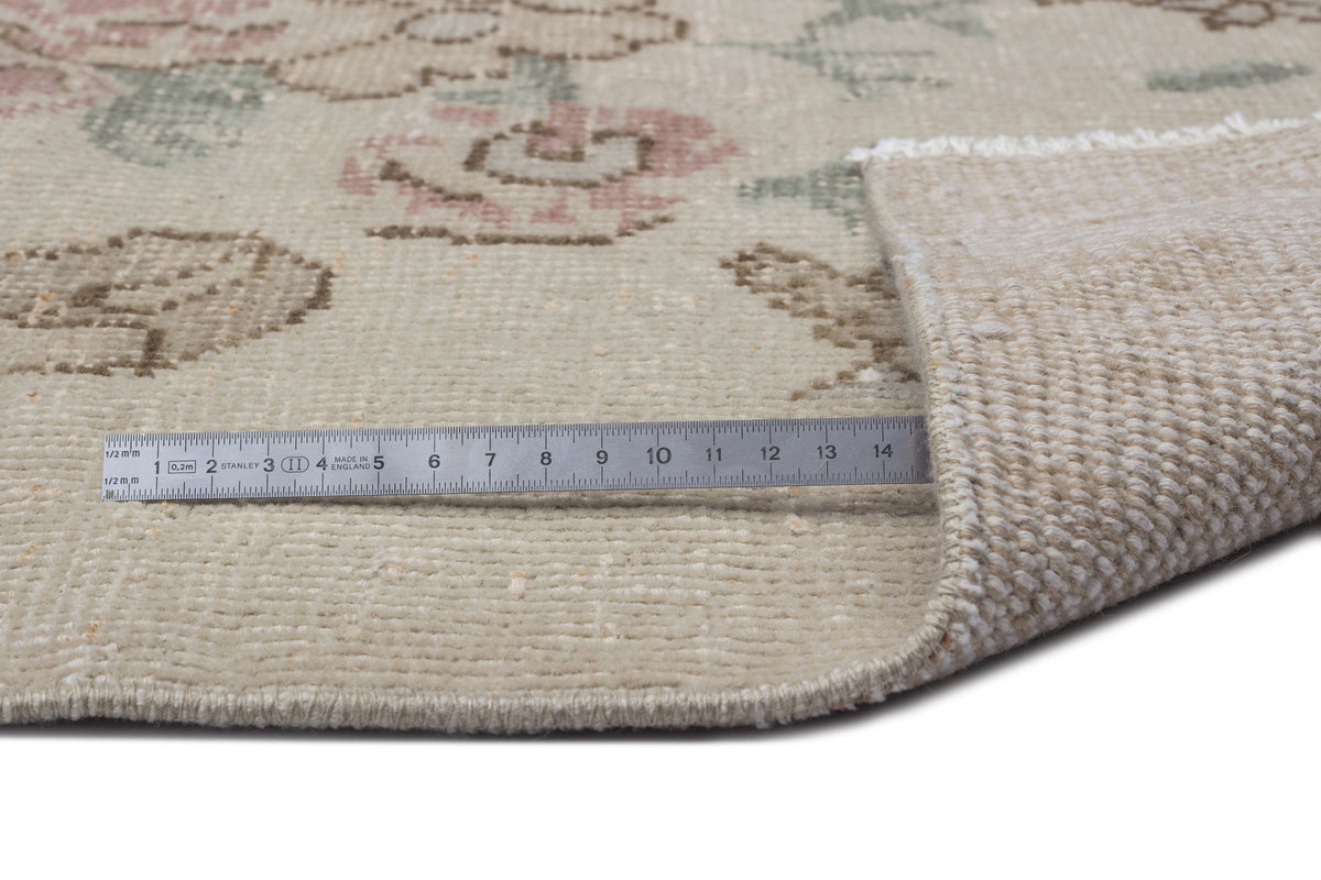 Beige rug with a faded floral pattern and a metal ruler showing pile thickness on a white background.