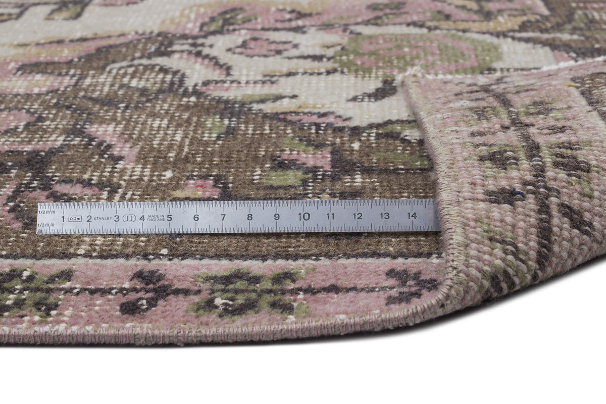 Distressed floral rug in brown, pink, and cream with a folded corner and a metal ruler for scale on a white background.