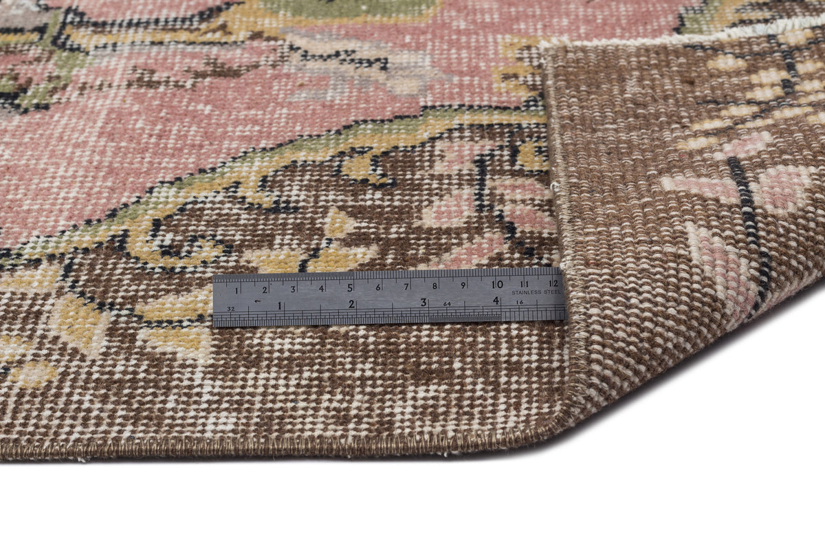 Distressed floral rug in pink, brown, and green with a folded corner and a metal ruler for scale on a white background.