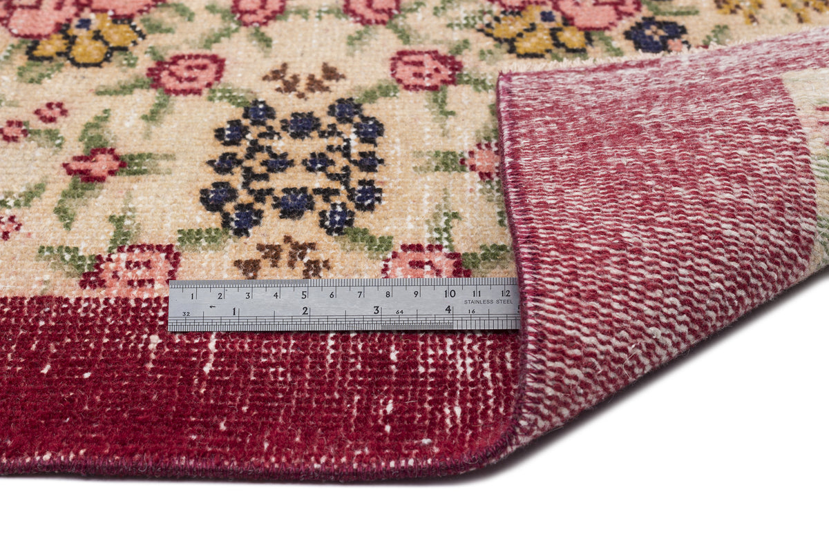 Beige rug with a floral pattern in pink, yellow, and blue, featuring a deep red border and a metal ruler for scale.