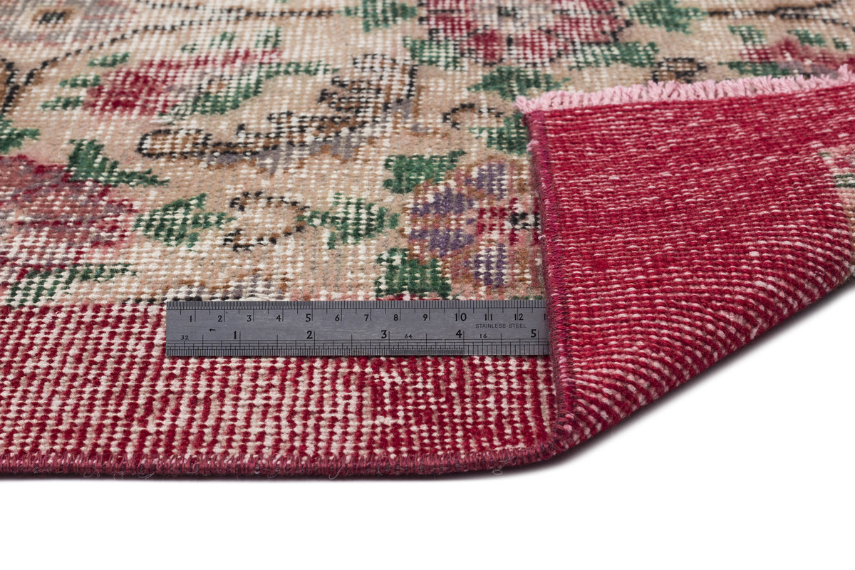 Beige rug with a distressed floral pattern in red, green, and purple, showing a folded corner and a metal ruler.