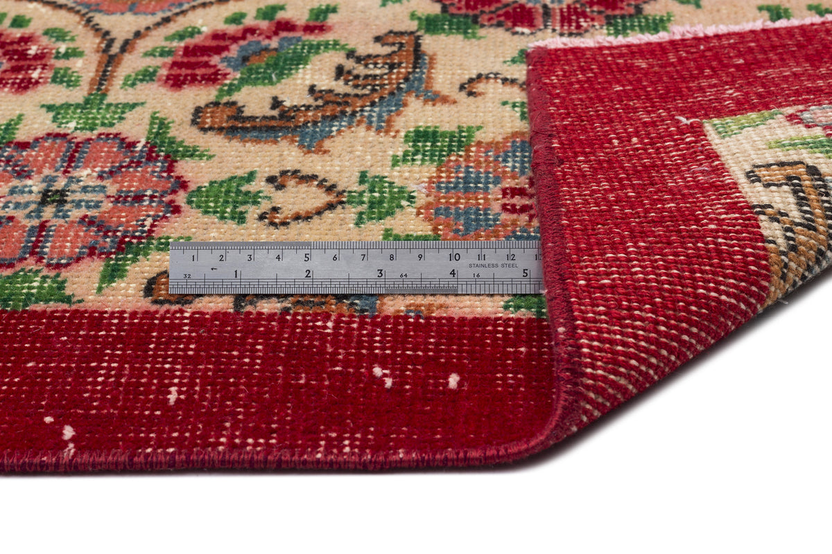 Beige rug with red, green, and blue floral pattern and a wide red border, shown with a metal ruler for scale.