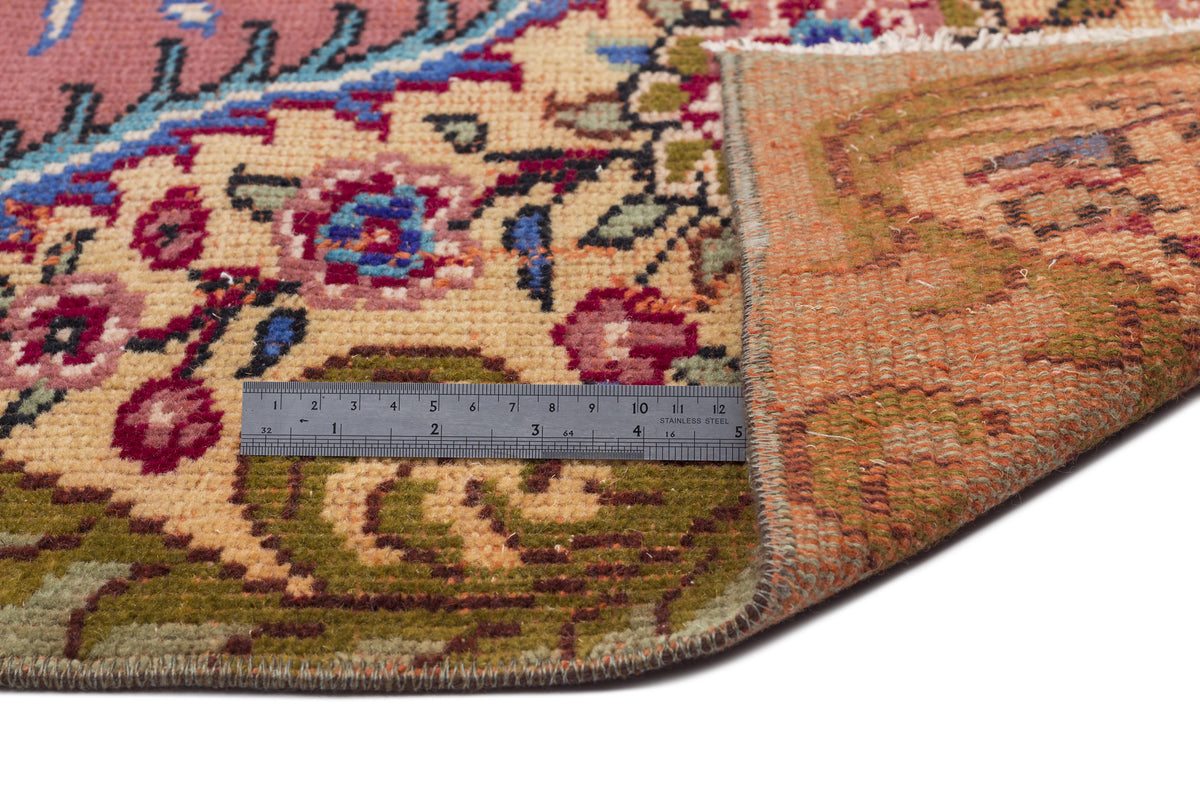Floral patterned rug with pink, beige, and green tones, shown folded to reveal the underside with a metal ruler for scale.