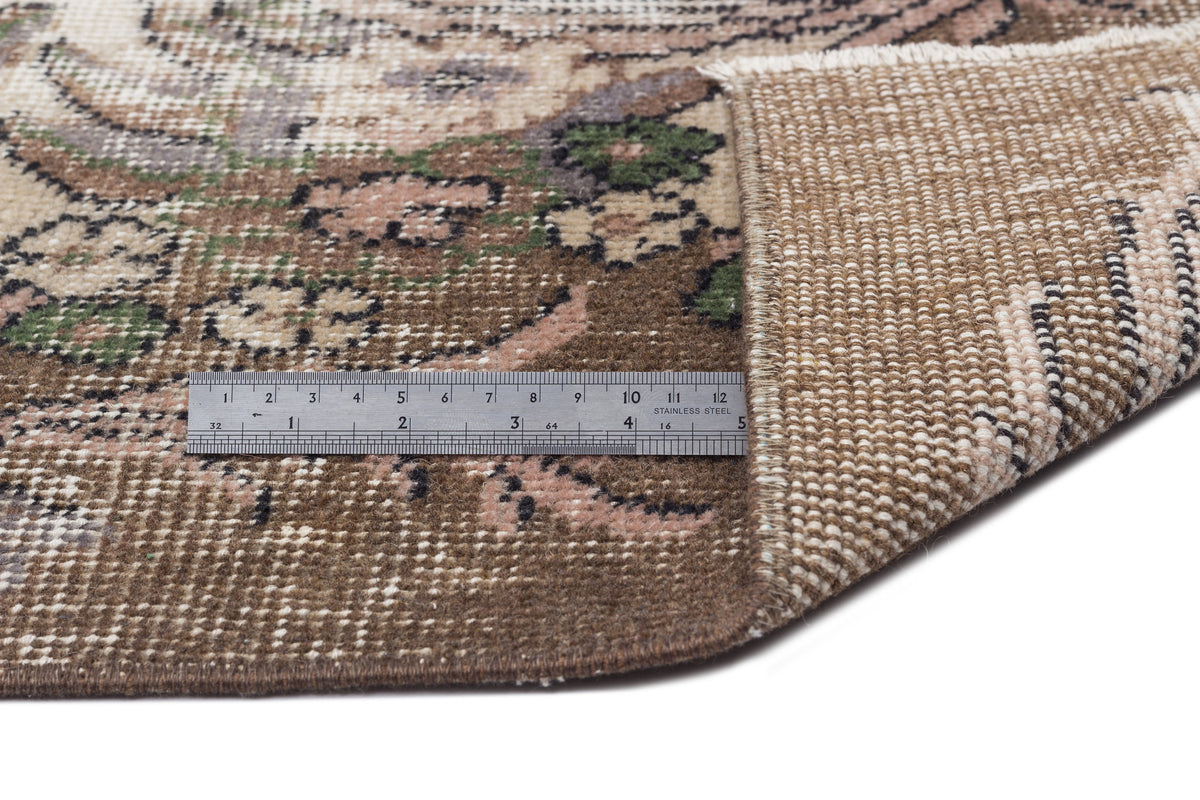 Brown, beige, and green floral patterned rug with a folded corner and a metal ruler for scale on a white background.