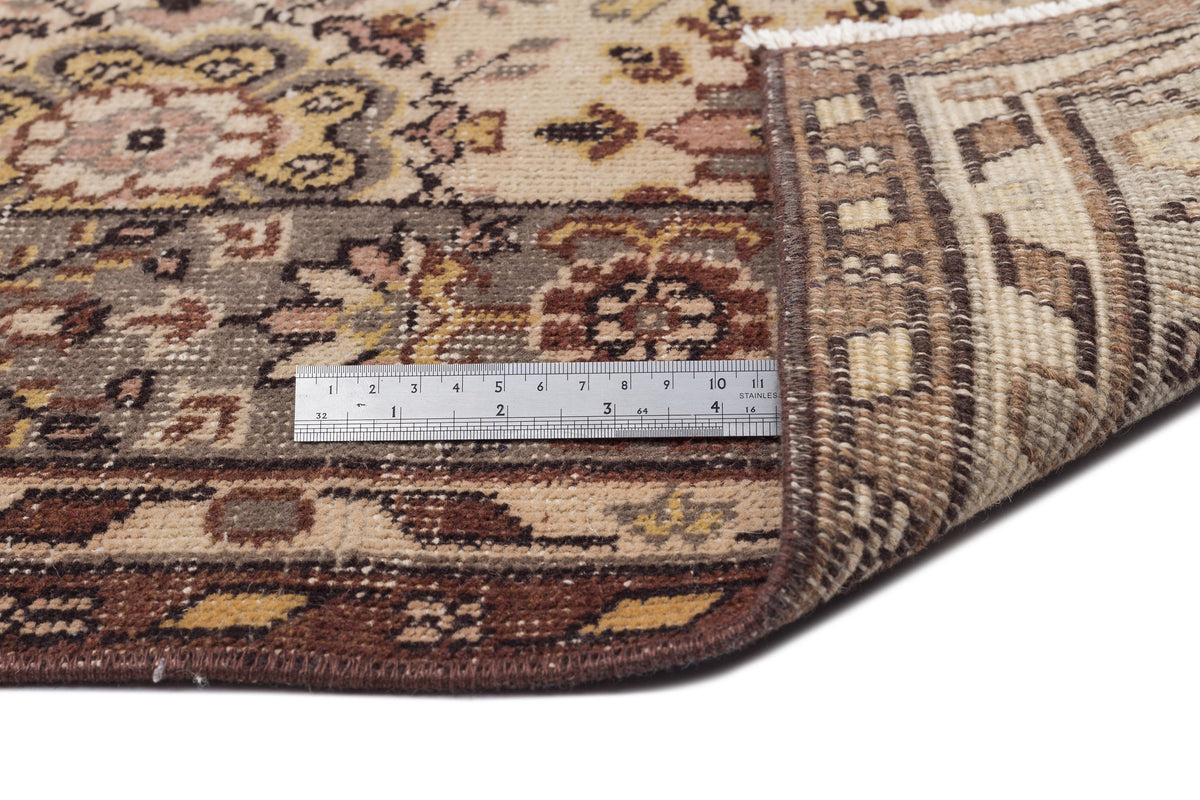 Beige and brown traditional floral rug with a folded corner and a metal ruler for scale on a white background.