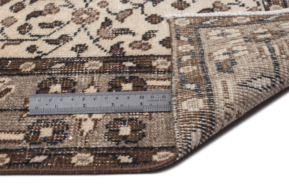 Beige and brown floral patterned rug with a folded corner and a metal ruler for scale on a white background.