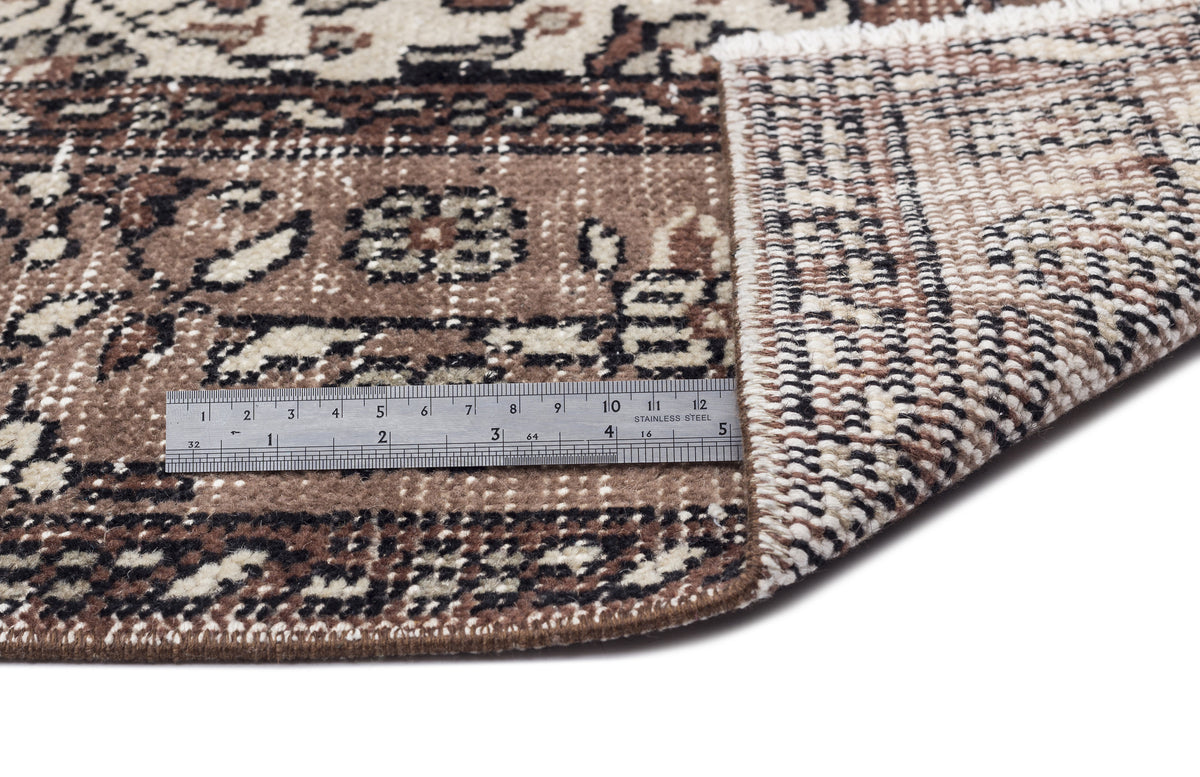 Brown, cream, and black traditional patterned rug with a folded corner showing the backing and a metal ruler for scale.