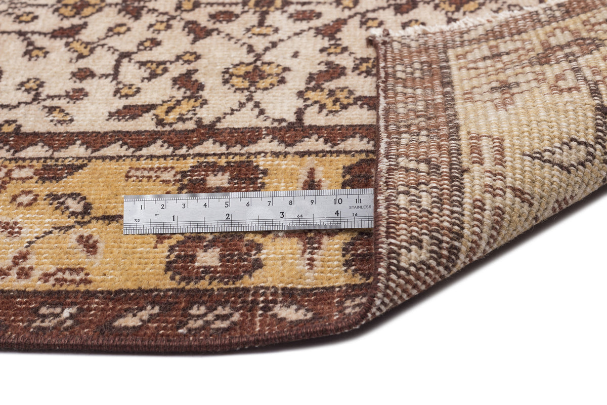 Beige and brown floral patterned rug with a folded corner and a metal ruler for scale on a white background.
