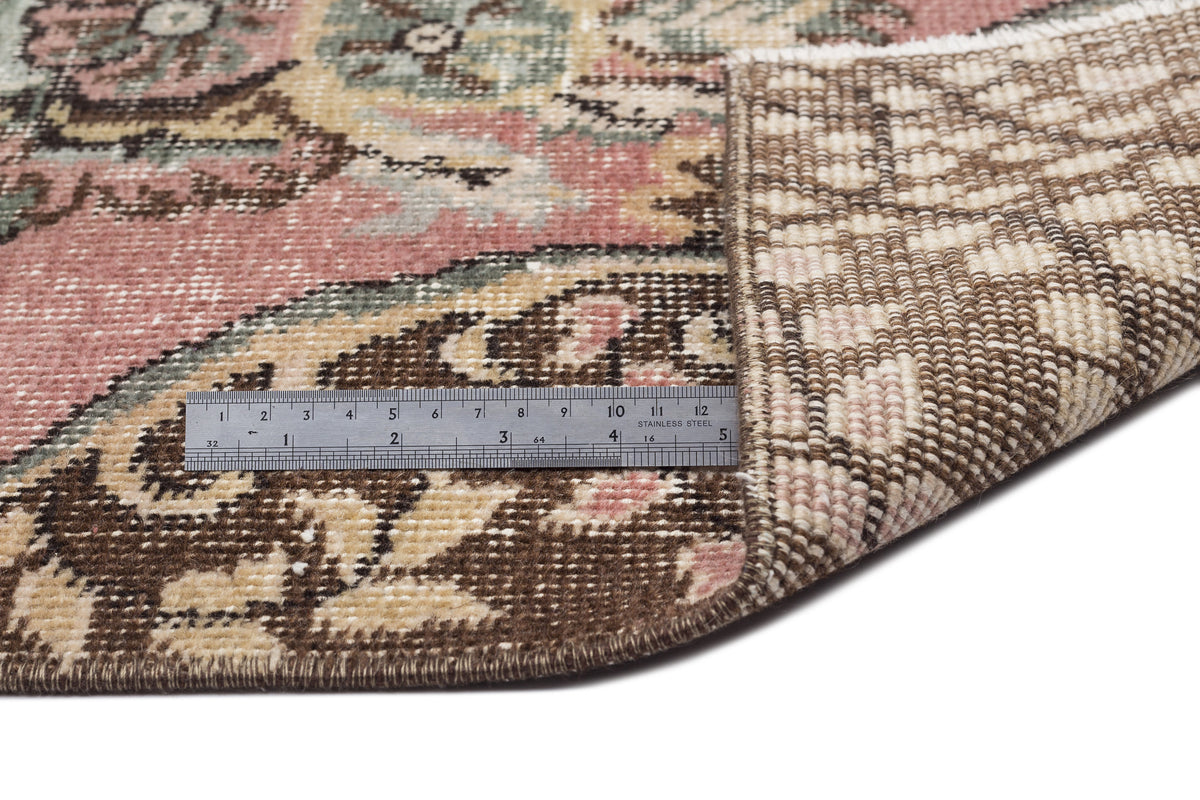 Pink, green, and brown floral medallion rug with a folded corner showing the woven backing and a metal ruler for scale.